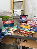 Photo showing full overview of all games and puzzles stacked on table including all titles visible and a sign noting ladder not for sale.