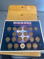Overall view of the commemorative coin set mounted on a card titled 'Man In Space' with background depicting space and stars. Coins are labeled with mission names from Mercury, Gemini, and Apollo programs. One coin slot is empty.