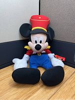 Front photo of a large Mickey Mouse plush dressed as a nutcracker soldier sitting on a wooden floor against a black and beige background. Ears show creases.