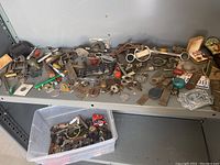 Wide view of assorted small antique collectibles including keys, mechanical parts, hardware, and coins arranged on a metal shelf and in a plastic container.