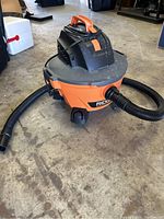 Orange and black Ridgid wet/dry vacuum with attached black flexible hose, on concrete floor showing general dust and wear on unit.