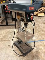 Front angle showing Ryobi drill press with control panel, worktable, spindle chuck with drill bit attached, and base.