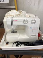 Front angle view of a white Singer sewing machine in its white carrying case, showing two control dials, thread spool, foot pedal, and some thread spools stored in the case.