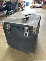 Closed heavy duty black travel case with two metal latches and top handle showing some wear and tear, placed on a concrete floor.