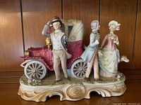Front and side views of a ceramic figurine featuring a vintage burgundy car and two men and two women in early 20th-century dress standing next to it on an ornate base.