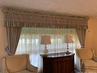 Window setup showing pleated valance, side drapes, sheer panels, a wooden cabinet between two chairs under the window and two table lamps on the cabinet.