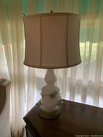 White ceramic table lamp with floral accents and beige fabric lampshade photographed indoors near window with sheer curtains.