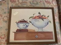 Framed watercolor style painting of two blue and white ceramic fruit bowls, one with peaches, one with strawberries, on a wooden surface with books beneath the strawberry bowl.