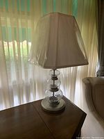 Glass table lamp with clear glass spherical base and white squared lampshade placed on a wooden table in front of curtains
