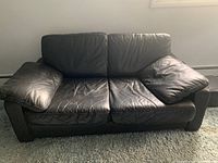 Front view of a black leather two-seat couch with some wrinkles in the leather cushions, placed on a carpeted floor against a white wall.