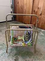 Gold-colored metal wire magazine rack holding a large cookbook, photographed against a wooden wall and concrete floor.