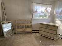 Wide shot showing dresser, headboard, and mirror leaning against the wall, all painted cream with green trim and floral designs.