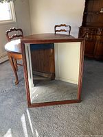 Rectangular mirror with wooden frame leaning against a table and wall, reflecting part of the room including a table and window.