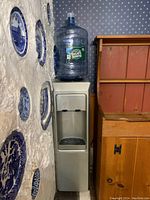 Photo of the Poland Springs water dispenser with attached water jug in corner between wall with decorative plates and wooden cabinet.