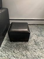 Small black leather ottoman shown on a carpeted floor next to a couch.