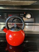 Red round tea kettle with black handle, black lid knob, and black spout cover on stovetop in front of an oven control panel.
