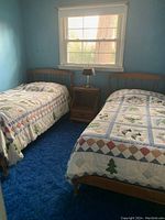 Two twin wooden beds positioned side by side in a blue carpeted room, each with nature-themed quilts and a wooden nightstand with a table lamp between them.