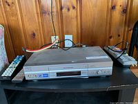 Silver Samsung VCR player with front RCA inputs and two remote controls on black table against wooden panel background.