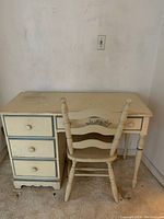 Full view of antique wooden writing desk and matching chair showing cream paint with blue trim and decorative accents. Three vertical drawers on one side and a center drawer with floral detail.