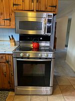 Photo shows Bosch stainless steel stove with glass cooktop and oven. Mounted above is a Bosch stainless steel microwave with digital controls. There is a red kettle on the stove. Wooden kitchen cabinets are on the sides.