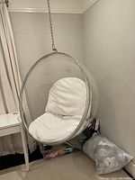 Photo of clear acrylic hanging egg chair with white cushion, hanging from metal chain attached to ceiling, placed in room corner near desk and curtains.