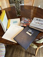 Four hardbound coffee table books on a wooden round table, including The Beatles Anthology, People Celebrates People 1974-1996, The Museum of Everything Volume 4, and The Appendix of Everything booklet.