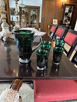 Green glass pitcher with pewter handle and base, six matching etched glasses arranged on a dark wood table.