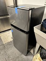 Full view of Insignia stainless steel apartment refrigerator with blue tape securing doors, showing top freezer and bottom refrigerator compartments.