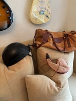 Set of three items on beige couch: black leather biker helmet, brown suede Bota bag, two-tone tan and brown leather satchel