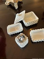 Four pieces of milk white hobnail glass arranged on a table: a lidded square box, two small rectangular bowls, and a lighter with matching hobnail glass outer case.