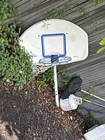 Outdoor basketball hoop leaning against wooden deck with visible dirt and wear, including backboard, blue hoop, and net. Propane tank near base not included.
