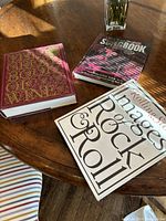 Photo of three large books on a round wooden table under sunlight: 'Rolling Stone Images of Rock & Roll', 'Songbook' music volume, and a red ornate book titled 'Wine'.