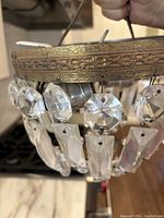 Close-up photo of vintage chandelier metal frame with hanging crystal prisms, showing etched floral pattern on circular rim and attached octagonal and rectangular crystals.