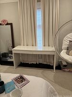 White desk positioned against wall with curtains, showing full front and top view in room context.