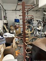 Full view of the tall wooden coat rack in a garage setting showing the vertical post with multiple sliding wooden rods extending horizontally for coat hanging, supported by a tripod base.