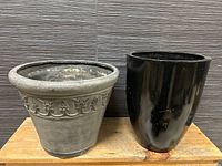 Front view of two large outdoor planters: gray planter with ornamental band on left, black glossy planter with surface scuffs on right.
