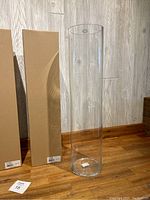 One clear tall cylindrical glass vase next to two cardboard boxes with labels on a wooden floor and wood panel wall background.