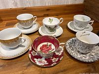 Photo shows six teacups and matching saucers on a wooden surface with detailed floral and decorative patterns.