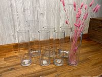 6 clear thick glass cylinder vases displayed upright on a wooden floor beside a wooden wall panel. One vase holds pink dried flower stems.