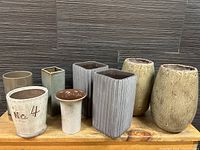 Wide shot showing all 8 ceramic planters and vases in various shapes and neutral tones placed on a wooden surface against a dark textured background.