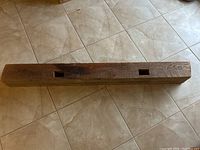 Long solid wood antique barn beam lying on tiled floor, showing full length and weathered surface with two square holes.