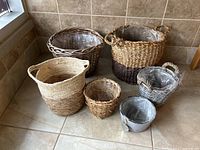 Group photo of 6 indoor planter baskets of varying sizes and woven materials, all lined with plastic. Placed on tiled floor close to window.
