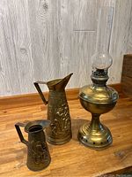 Full view of brass oil lamp with glass chimney and two brass jugs on wooden surface.