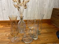 All six etched glass vases of varying shapes and sizes arranged on a wood surface against a light wood panel background, showing etched floral and leaf patterns.