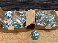 Two cardboard boxes containing multiple turquoise and white ceramic drawer knobs with brass fixtures. One knob placed outside boxes for display.