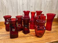 Ten assorted red glass vases arranged on wooden surface against a light wood background, various shapes and sizes, some with black leather straps and metal heart pendants.