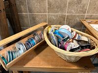 Wide view showing a wooden ribbon roll holder filled with various satin ribbon spools and a basket beside it holding additional ribbon spools.