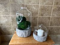 Photo showing both candle holders: one larger with moss balls, one smaller with white candle and decorative spheres under glass hurricanes, both with cement bases shaped like tree trunks.