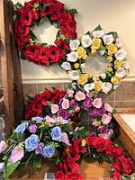 Photo showing five different silk flower wreaths including red poppy, white and yellow roses, pink and purple roses, and blue roses arranged on a shelf.