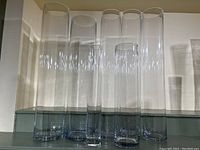 Set of 7 tall cylinder glass vases of varying heights photographed on a shelf, clear glass, smooth surface.